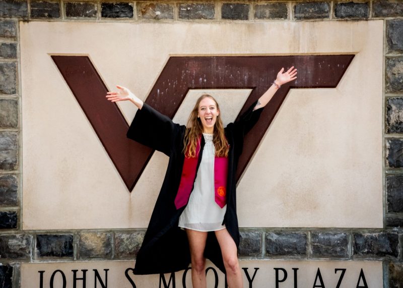 Female student smiles big with arms open in front of stone VT wall in graduation attire.